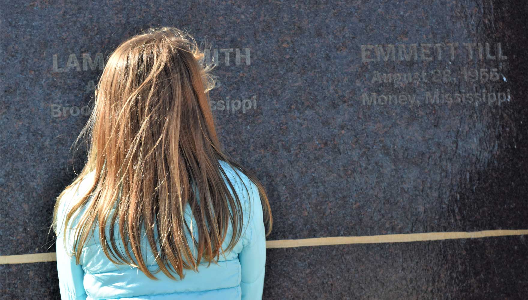 A young girl with blonde hair contemplates the monument at the Peace and Justice Memorial Center, which memorializes people who were victims of racial violence during the 1950s, including Emmett Till, whose name and the date and place of his murder are inscribed on the monument.