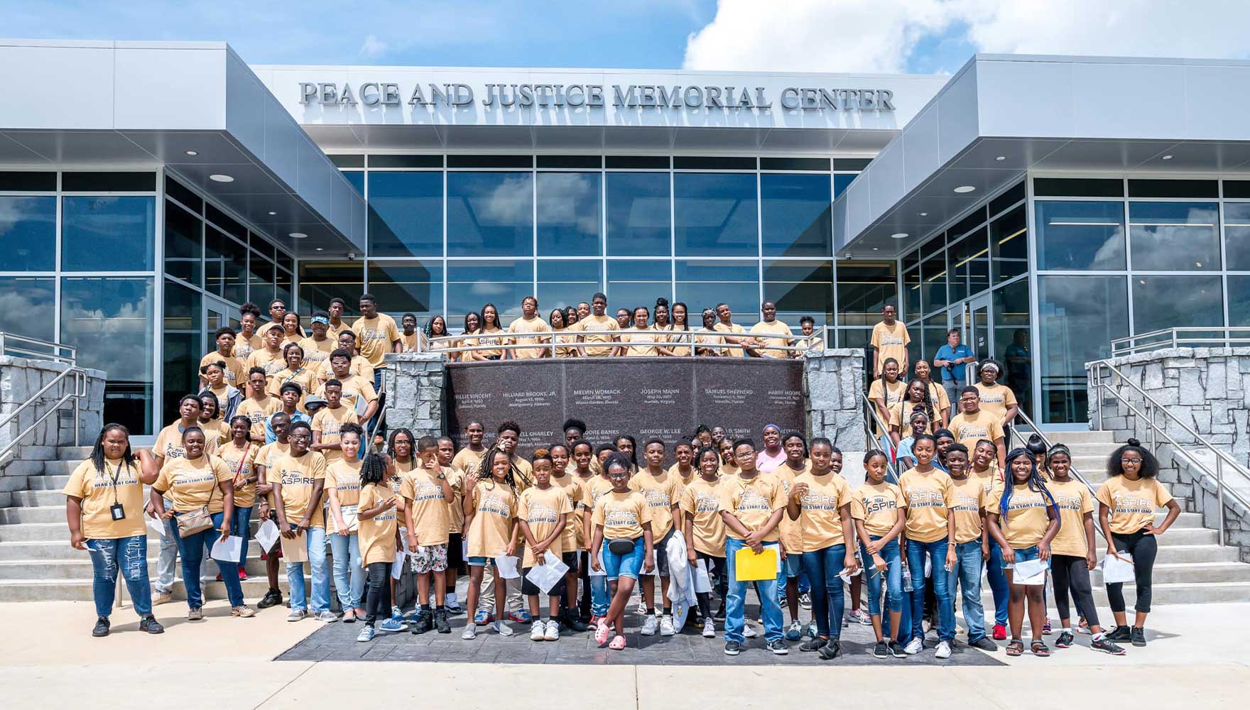 A large group of high school students in matching t-shirts pose outside the Peace and Justice Memorial Center.