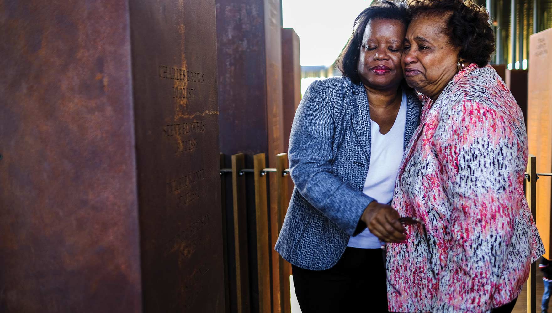 Two women embrace tearfully in front of a monument on which the name of their ancestor is inscribed at the National Memorial for Peace and Justice.